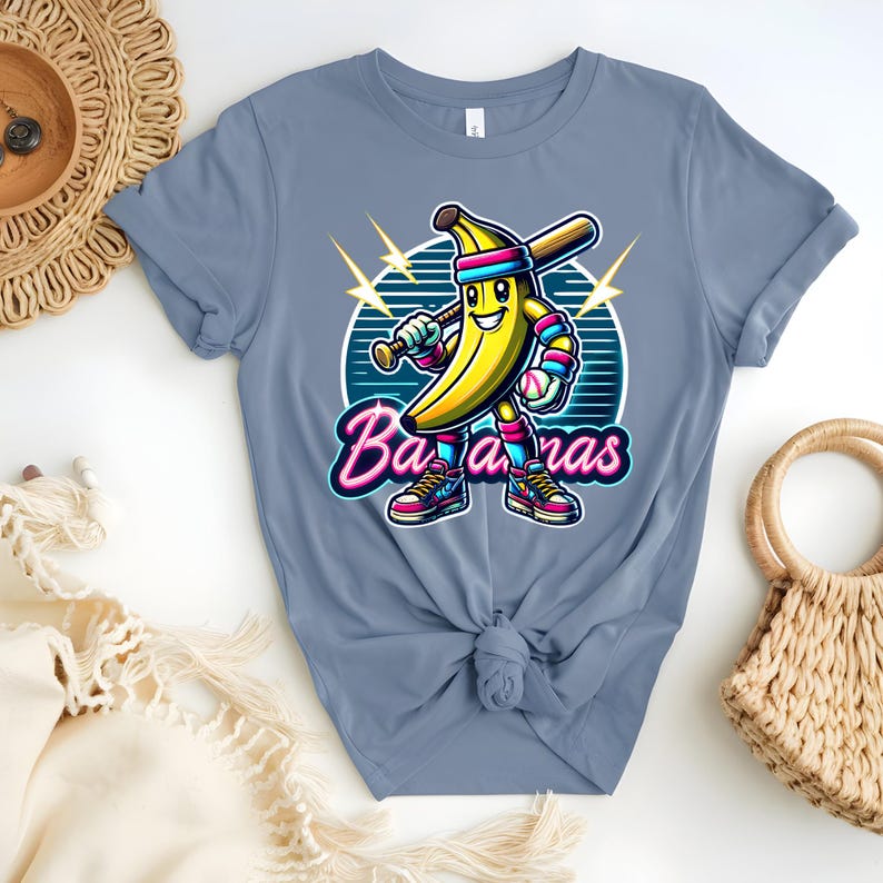 K&ouml;nnte beinhalten: Ein hellblaues T-Shirt mit einer Cartoon-Banane, die einen Baseballschl&auml;ger h&auml;lt und eine Baseballkappe tr&auml;gt. Die Banane tr&auml;gt Turnschuhe und hat ein L&auml;cheln im Gesicht. Der Text "Bananas" ist in Neon-Pink und Blau geschrieben.