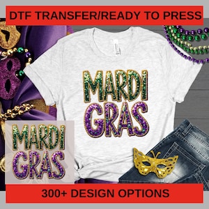 Puede incluir: Una camiseta blanca con las palabras "MARDI GRAS" en purpurina verde y morada. La imagen incluye una máscara dorada, cuentas y el texto "300+ DESIGN OPTIONS" en una pancarta roja, lo que sugiere una selección de diseños.