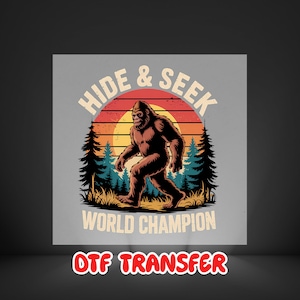 May include: A graphic design featuring a brown Bigfoot walking in front of a sunset with the text "HIDE & SEEK WORLD CHAMPION". The design includes pine trees and grass. The words "DTF TRANSFER" are at the bottom.