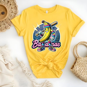 K&ouml;nnte beinhalten: Gelbes T-Shirt mit einer Cartoon-Banane, die ein Baseball-Uniform tr&auml;gt und einen Schl&auml;ger h&auml;lt. Der Text "Bananas" ist in rosa und blauen Buchstaben geschrieben.