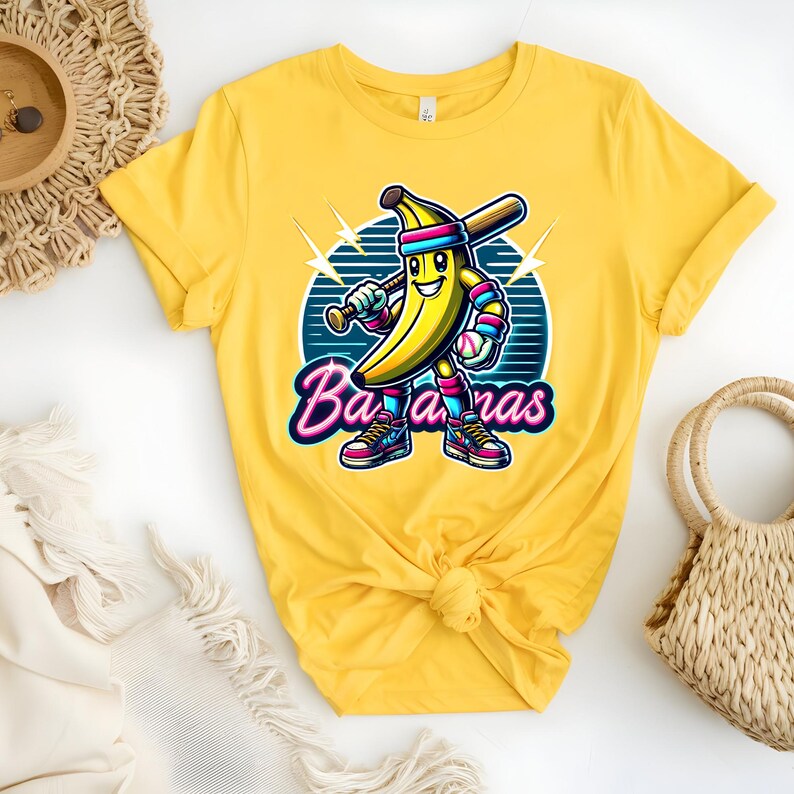 K&ouml;nnte beinhalten: Gelbes T-Shirt mit einer Cartoon-Banane, die ein Baseball-Uniform tr&auml;gt und einen Schl&auml;ger h&auml;lt. Der Text "Bananas" ist in rosa und blauen Buchstaben geschrieben.