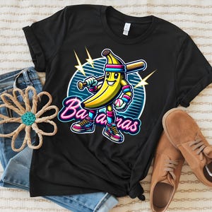 K&ouml;nnte beinhalten: Schwarzes T-Shirt mit einer Cartoon-Bananenfigur, die einen Baseballschl&auml;ger und einen Baseball h&auml;lt. Die Banane tr&auml;gt einen rosa-blauen Hut und Turnschuhe. Der Text "Bananas" ist in Neon-Pink und Blau.