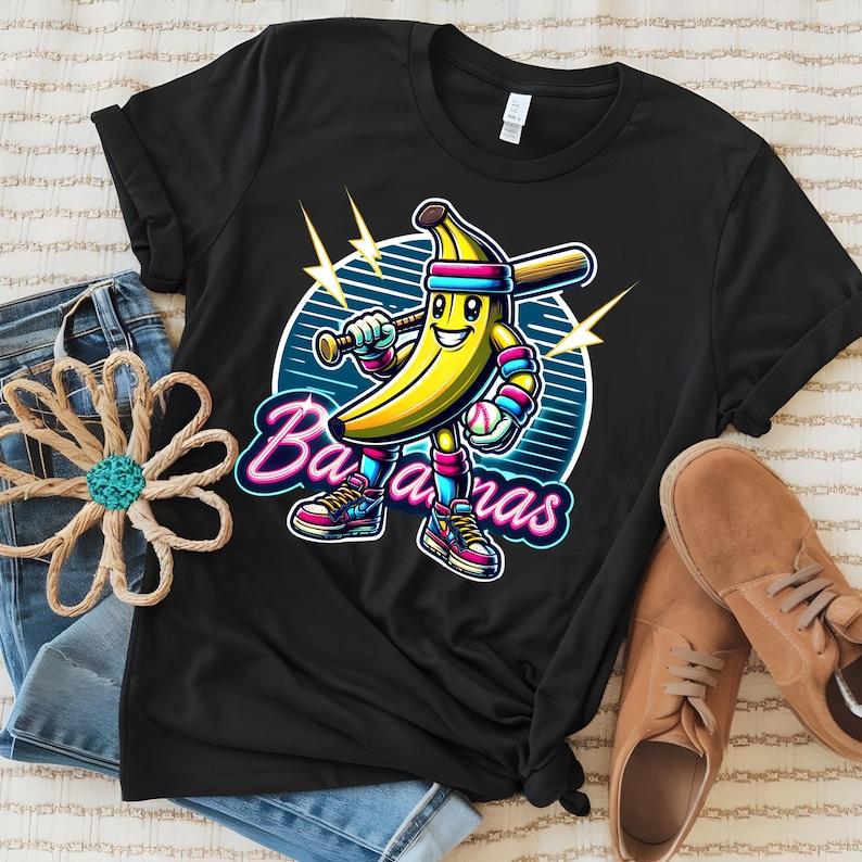 K&ouml;nnte beinhalten: Schwarzes T-Shirt mit einer Cartoon-Bananenfigur, die einen Baseballschl&auml;ger und einen Baseball h&auml;lt. Die Banane tr&auml;gt einen rosa-blauen Hut und Turnschuhe. Der Text "Bananas" ist in Neon-Pink und Blau.