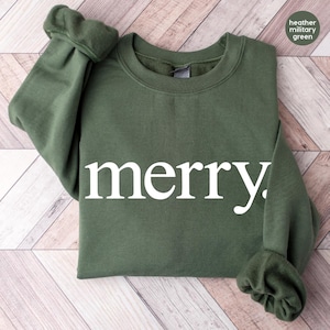 Puede incluir: Una sudadera verde militar jaspeada con la palabra "merry" en letras blancas. La sudadera tiene cuello redondo y mangas largas con puños acanalados. La prenda está doblada sobre una superficie de vetas de madera clara.