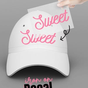 K&ouml;nnte beinhalten: Wei&szlig;e Baseballkappe mit einem rosafarbenen B&uuml;gelbild, auf dem "Sweet Sweet" steht. Das B&uuml;gelbild wird mit einem Blatt durchsichtigem Papier auf die Kappe aufgebracht.