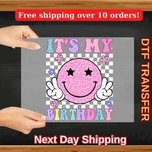 May include: A DTF transfer sheet featuring a pink smiley face with black star eyes and peace sign hands. The words "IT'S MY BIRTHDAY" are in colorful, patterned letters. The background is a checkered pattern.