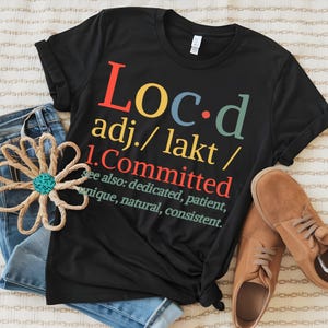 Könnte beinhalten: Schwarzes T-Shirt mit dem Wort "Loc.d" in bunten Buchstaben. Das Shirt enthält auch die Definition des Wortes "Committed". Das Shirt wird mit blauen Jeans und braunen Schuhen kombiniert.