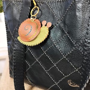 Könnte beinhalten: Schwarze Lederhandtasche mit Steppmuster und Ziernähten. Ein dekorativer Schneckenanhänger mit orangefarbenen, gelben und rosa Akzenten hängt am Griff. Die Tasche hat einen langen Riemen und ein kleines Logo.