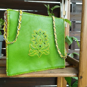 Könnte beinhalten: Eine hellgrüne Ledertasche mit einem dekorativen gelben Blumendesign. Die Tasche hat einen Kettenriemen mit gelben Akzenten und gelben Nähten. Die Tasche hat eine rechteckige Form.