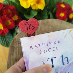 Könnte beinhalten: Ein roter herzförmiger Lesezeichen mit dem Buchstaben "M" und einem kleinen Herzdetail, das in einem Buch platziert ist. Der Buchtitel ist teilweise sichtbar, mit dem Namen des Autors "Kathinka Engel". Gelbe und rote Blumen sind im Hintergrund.