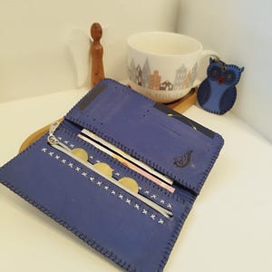 Könnte beinhalten: Offene blaue Ledergeldbörse mit silbernem Reißverschluss und weißen Nähten. Die Geldbörse enthält Münzen und Banknoten. Ein eulenformiger Schlüsselanhänger und eine Tasse mit einer Stadtszene sind ebenfalls zu sehen.