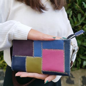 Op de afbeelding: Een rechthoekige patchwork leren clutch. Het heeft vierkanten in verschillende kleuren, waaronder paars, blauw, roze en groen, aan elkaar gestikt met witte draad. Het heeft een bruine riem en een kleine polsband.