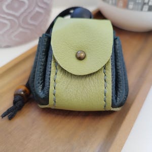 Könnte beinhalten: Kleine, handgefertigte Ledertasche in Grün- und Dunkelblautönen. Die Tasche hat einen Klappenverschluss mit Messingknopf und einen Kordelzug mit Holzperle. Die Tasche liegt auf einem Holztablett.