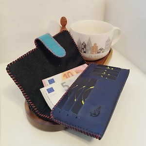Könnte beinhalten: Eine schwarze Geldbörse mit türkisblauem Verschluss und roten Nähten, gefüllt mit Euro-Banknoten. Ein blaues Kartenetui mit roten Nähten daneben. Eine weiße Tasse mit Stadtmotiv und dem Text "ZU HAUSE" im Hintergrund.