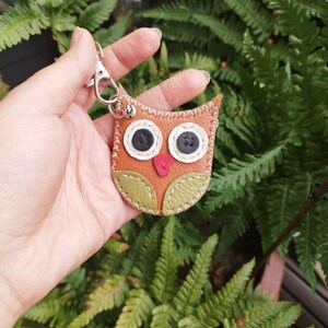 Könnte beinhalten: Ein brauner Leder-Eulen-Schlüsselanhänger mit grünen Akzenten und schwarzen Augen. Die Eule hat ein gesticktes Design und einen roten Schnabel.