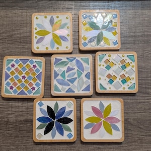 May include: Seven square coasters with mosaic tile designs. Each coaster has a bamboo frame and a unique pattern of colourful glass tiles, including floral and geometric arrangements. The colour palette includes blues, greens, yellows, and pinks.
