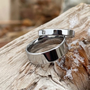 May include: Two polished silver-toned metal rings with a smooth, reflective surface. The rings feature a simple, flat band design with a high-shine finish. One ring is positioned slightly behind and above the other, showcasing their metallic luster.