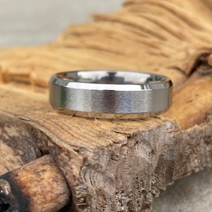 May include: A silver-colored ring with a brushed metal finish. The ring is set on a piece of rough-hewn wood, showcasing its texture and design. The ring's band is approximately 0.25 inches wide. The ring is a piece of jewelry.