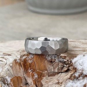 May include: A silver hammered wedding band with a textured, geometric design. The band is sitting on a piece of weathered wood.