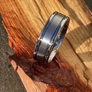 May include: A silver-colored ring with a brushed center band and polished edges. The ring is set against a backdrop of natural wood with rich brown and reddish hues, creating a contrast of textures and colors.