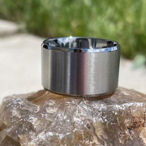 May include: A silver ring with a brushed finish and a beveled edge. The ring is sitting on a light brown rock.