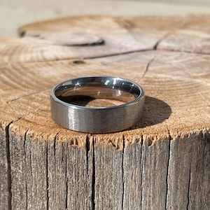 May include: A metallic ring with a brushed finish and a flat profile. The band features a smooth, matte-textured outer surface with a polished, reflective inner edge, showcasing a neutral gray tone that catches the light across its circular form.