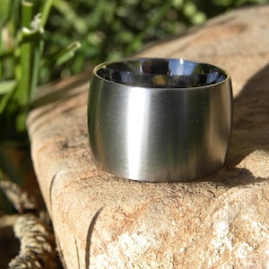 May include: A wide, brushed stainless steel ring with a cylindrical shape. The ring sits on a textured, light brown stone, with green foliage in the background. The ring's interior is dark.