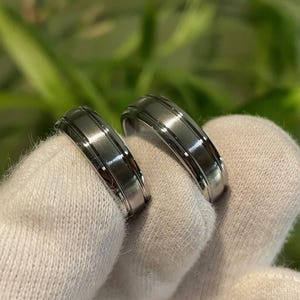 May include: Two matching silver-toned metal rings with a polished finish. Each ring features a central band with a brushed texture, flanked by two thin, raised, and polished grooves that run along the entire circumference of the circular band.