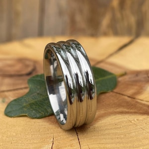 May include: A shiny, silver-colored ring with a ribbed design. The ring is set on a wooden surface with a green leaf. The ring's surface reflects the surrounding environment.