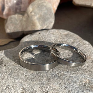 May include: Two silver wedding bands, one slightly wider than the other, are placed on a gray rock.