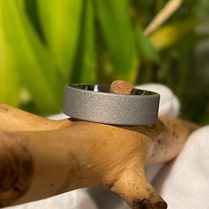 May include: A silver ring with a textured finish. The ring is displayed on a piece of wood. The ring has a flat band design. The ring is a piece of jewellery.