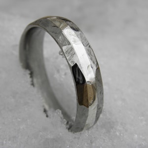 以下が含まれることがあります： 中央に磨かれたストライプのある、銀製のハンマーで叩かれた結婚指輪。指輪は雪の表面に置かれています。