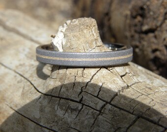 May include: A close-up of a ring with a gray exterior and a gold-colored center band. The ring is set against a weathered, light brown wooden surface, with natural light illuminating the details of the ring's texture and design.