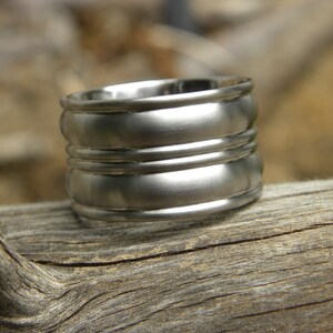 May include: A wide silver-toned metal ring featuring a smooth, matte finish with two prominent horizontal raised bands encircling the center. The band has a thick, substantial profile with rounded edges along the top and bottom rims.