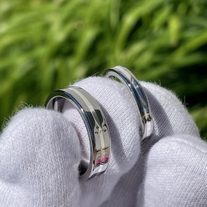 May include: Two silver rings with a textured design are displayed. The rings are held against a white fabric background, with a blurred green background. The rings have a polished, reflective surface.