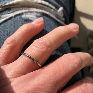 May include: A silver band ring is worn on a finger. The ring is thin and has a smooth, polished surface. The ring is a simple, elegant piece of jewellery. The background shows a person wearing blue jeans.
