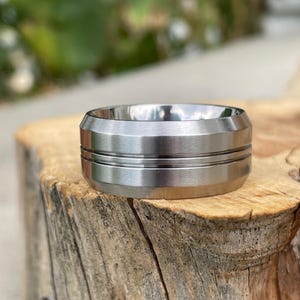 May include: A silver-toned metal ring with a brushed finish, featuring a wide band with beveled edges and two thin, horizontal grooves running around the center of the exterior surface, creating a distinct layered appearance on the metal band.