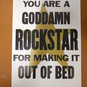 May include: A white poster with black text and a gold star graphic. The text reads: "YOU ARE A GODDAMN ROCKSTAR FOR MAKING IT OUT OF BED."