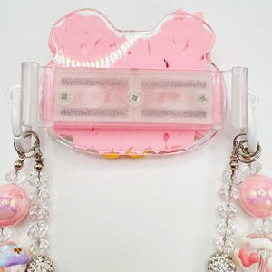 以下が含まれることがあります： ピンク色のリボン型のアクセサリーホルダー。透明なプラスチッククリップとビーズネックレスが付いています。ネックレスはピンク、クリア、シルバーのビーズと装飾的なチャームが特徴です。ホルダーは白い表面に取り付けられています。