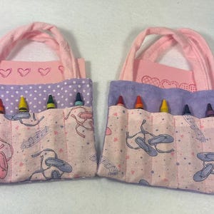 May include: Two pink fabric bags with purple trim and a pink and white patterned lining. Each bag has six slots for crayons and a small pocket for a notepad. The bags have pink handles.