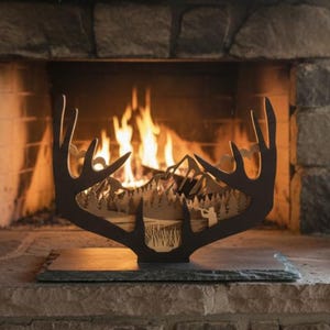 May include: A decorative metal antler silhouette with a mountain scene cut out, placed on a slate base in front of a fireplace. The flames of the fire are visible behind the silhouette. A painting and a glass terrarium are also visible.