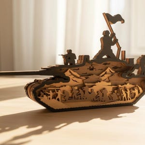 May include: A detailed wooden tank model with cut-out silhouettes of soldiers, a helicopter, and mountainous terrain. The tank features a long barrel and figures holding a flag. The model is made of light brown wood and sits on a wooden surface.