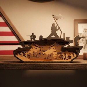 May include: A layered wooden tank sculpture with soldiers and a flag. The tank is brown with detailed cutouts depicting a battle scene. An American flag and a framed photograph are in the background.