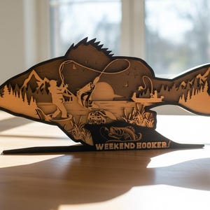 May include: A layered wooden fish-shaped art piece with a fishing scene. The design features a fisherman, a boat, and a fish jumping out of the water. The text "WEEKEND HOOKER" is at the base.
