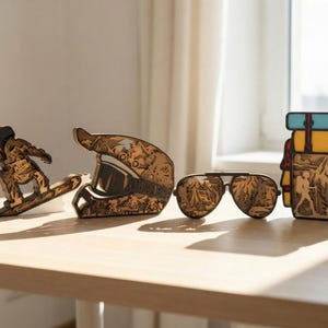 May include: Wooden decorative items including a snowboarder, a motorcycle helmet, aviator sunglasses, and a backpack. The items are made of layered wood, with intricate details. The sunglasses have a mountain scene reflected in the lenses.