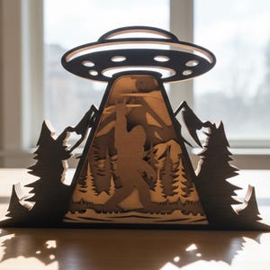 May include: A wooden layered art piece depicting a UFO hovering over a mountain scene with a Bigfoot figure. The artwork is in shades of brown and black, with trees and mountains. The UFO has a circular design.