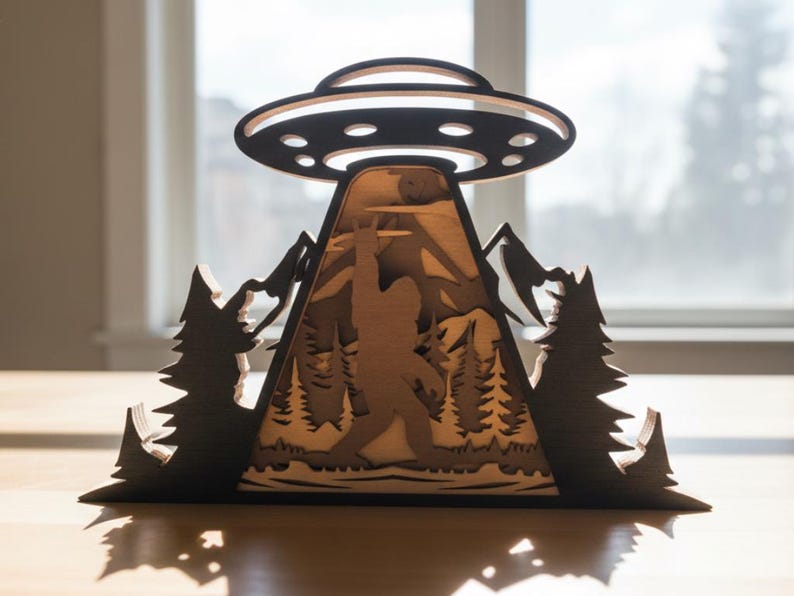 May include: A wooden layered art piece depicting a UFO hovering over a mountain scene with a Bigfoot figure. The artwork is in shades of brown and black, with trees and mountains. The UFO has a circular design.