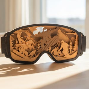 May include: Wooden decorative goggles with a layered design depicting a snowboarding scene. The goggles are black with a light brown interior, featuring silhouettes of skiers, trees, and mountains. The goggles are resting on a light-colored surface.
