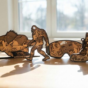 May include: Wooden laser-cut art pieces depicting outdoor themes. Includes a fish with a fisherman, Bigfoot, a pair of goggles with a mountain scene, and a hiking boot. The pieces are brown and stand on a light-colored surface.