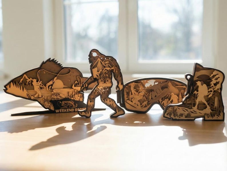 May include: Wooden laser-cut art pieces depicting outdoor themes. Includes a fish with a fisherman, Bigfoot, a pair of goggles with a mountain scene, and a hiking boot. The pieces are brown and stand on a light-colored surface.